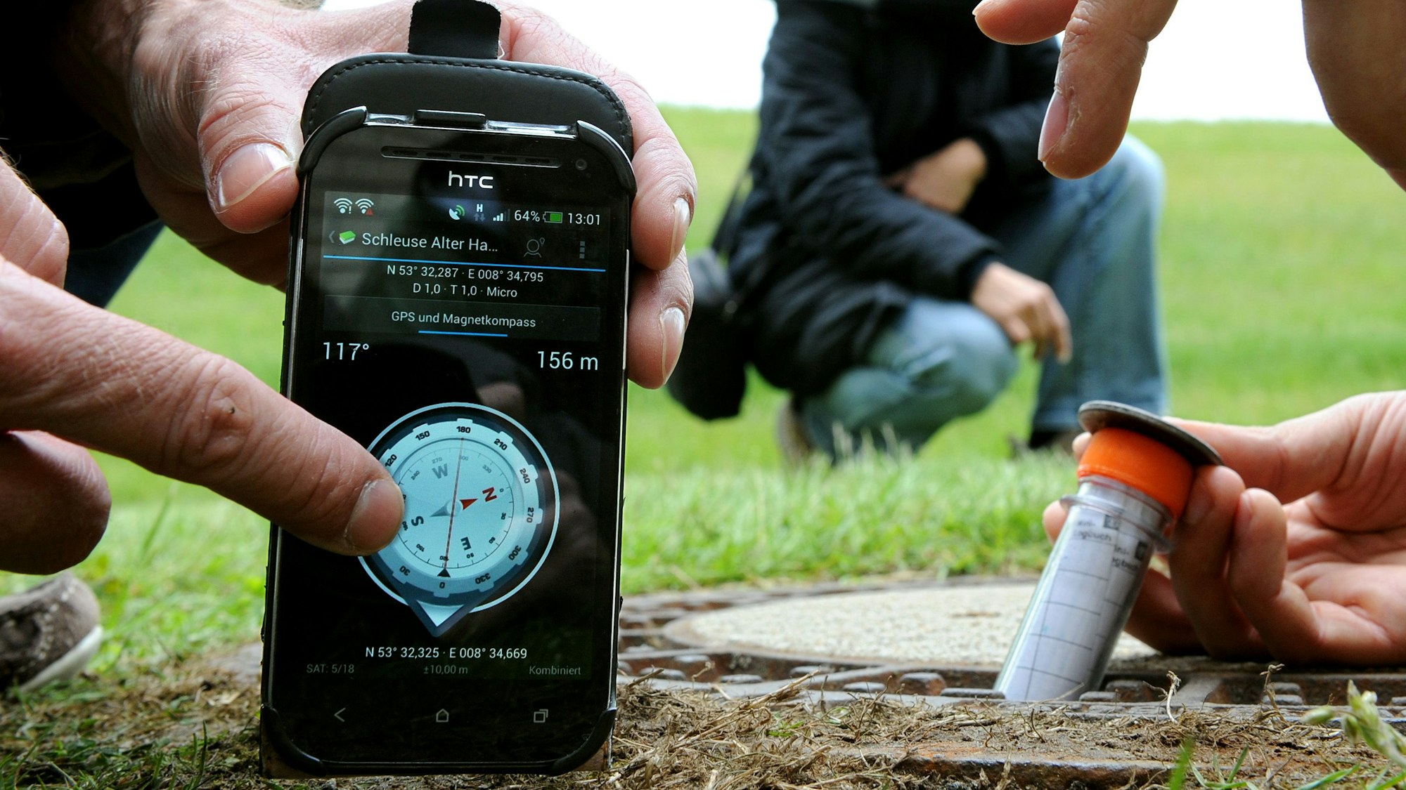 Auf einem Handy ist ein digitaler Kompass zu sehen, der GPS-Koordinaten anzeigt. Daneben nimmt eine Person ein Röhrchen mit einem Geo-Cache aus dem Boden.