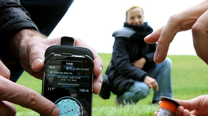 Auf einem Handy ist ein digitaler Kompass zu sehen, der GPS-Koordinaten anzeigt. Daneben nimmt eine Person ein Röhrchen mit einem Geo-Cache aus dem Boden.