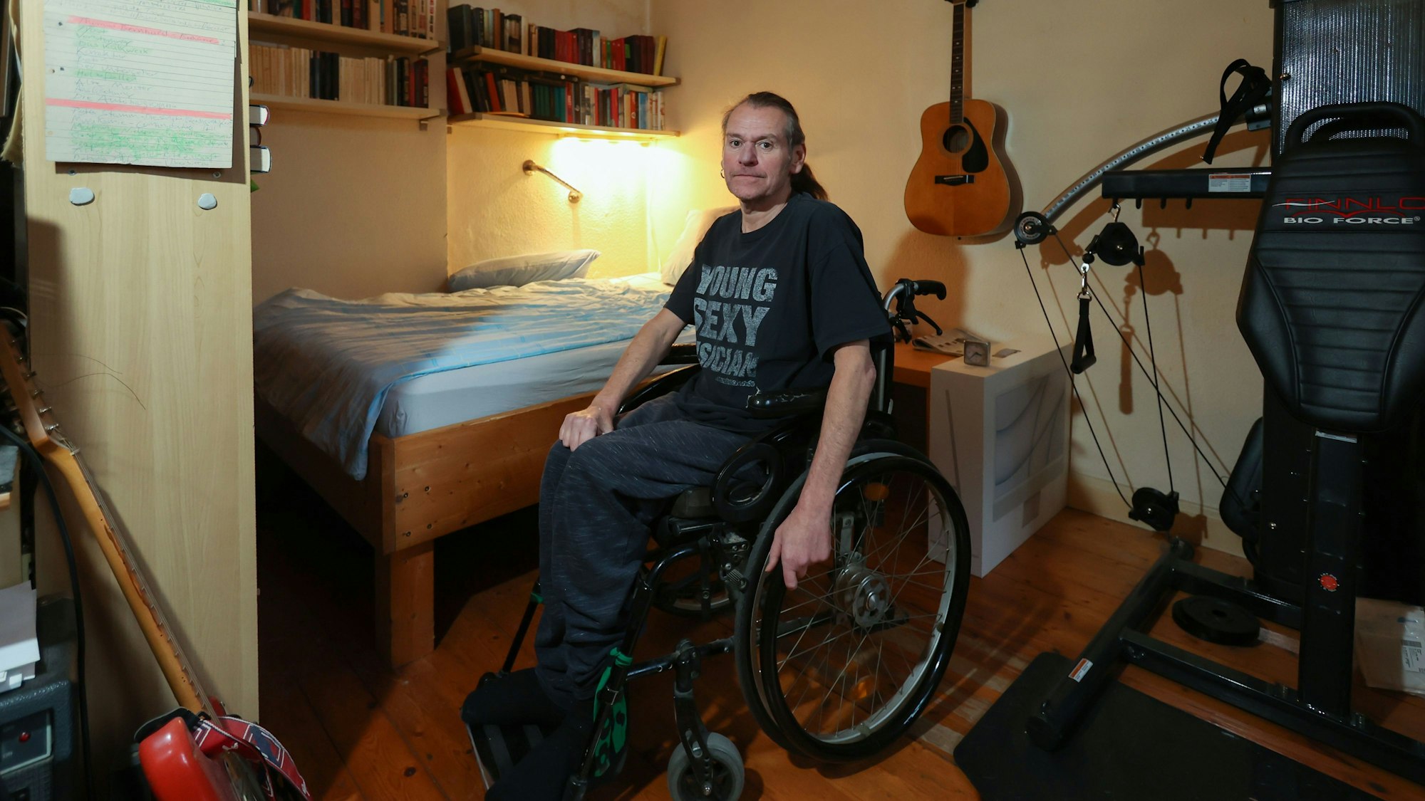 Ralf Degen sitzt im Rollstuhl. Hinter ihm ist sein Bett zu sehen, rechts von ihm ein Fitnessgerät. Er trägt ein T-Shirt mit der Aufschrift Young, Sexy, Muscican.