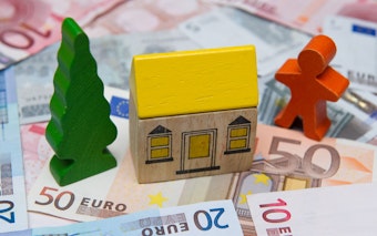 Man sieht ein Bauklotz-Haus, das auf Geldscheinen steht. Rechts neben dem Haus steht eine Holzfigur, links ein Holzbaum.