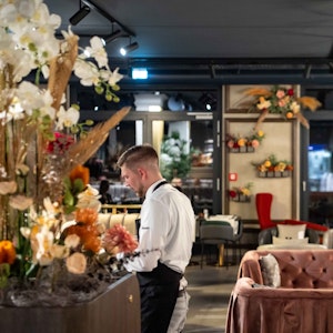 Ein Restaurant ist mit zahlreichen Blumen dekoriert, ein Kellner steht an einem Tisch.