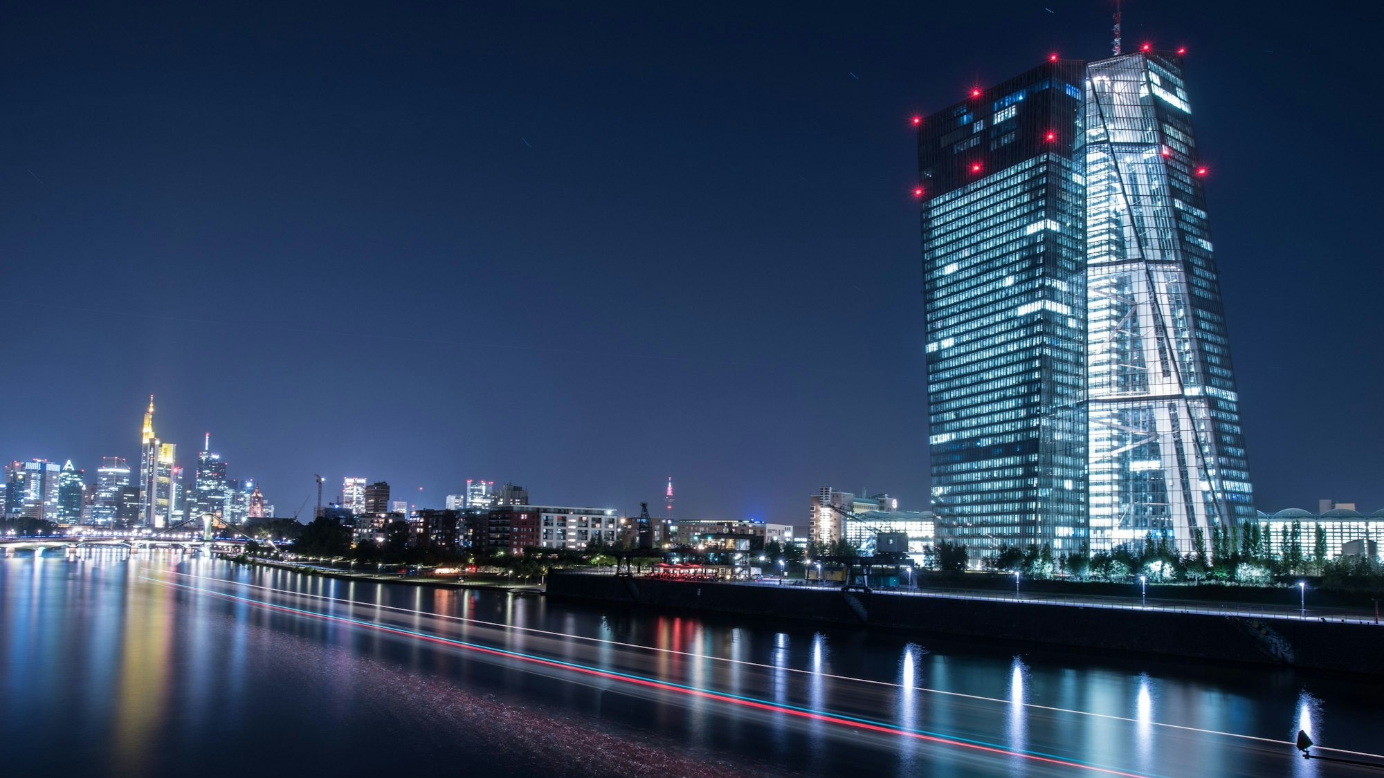 Die Lichter der Europäischen Zentralbank (EZB) leuchten am in Frankfurt am Main, während ein auf dem Main vorbeifahrendes Schiff Lichtspuren durch die Aufnahme zieh