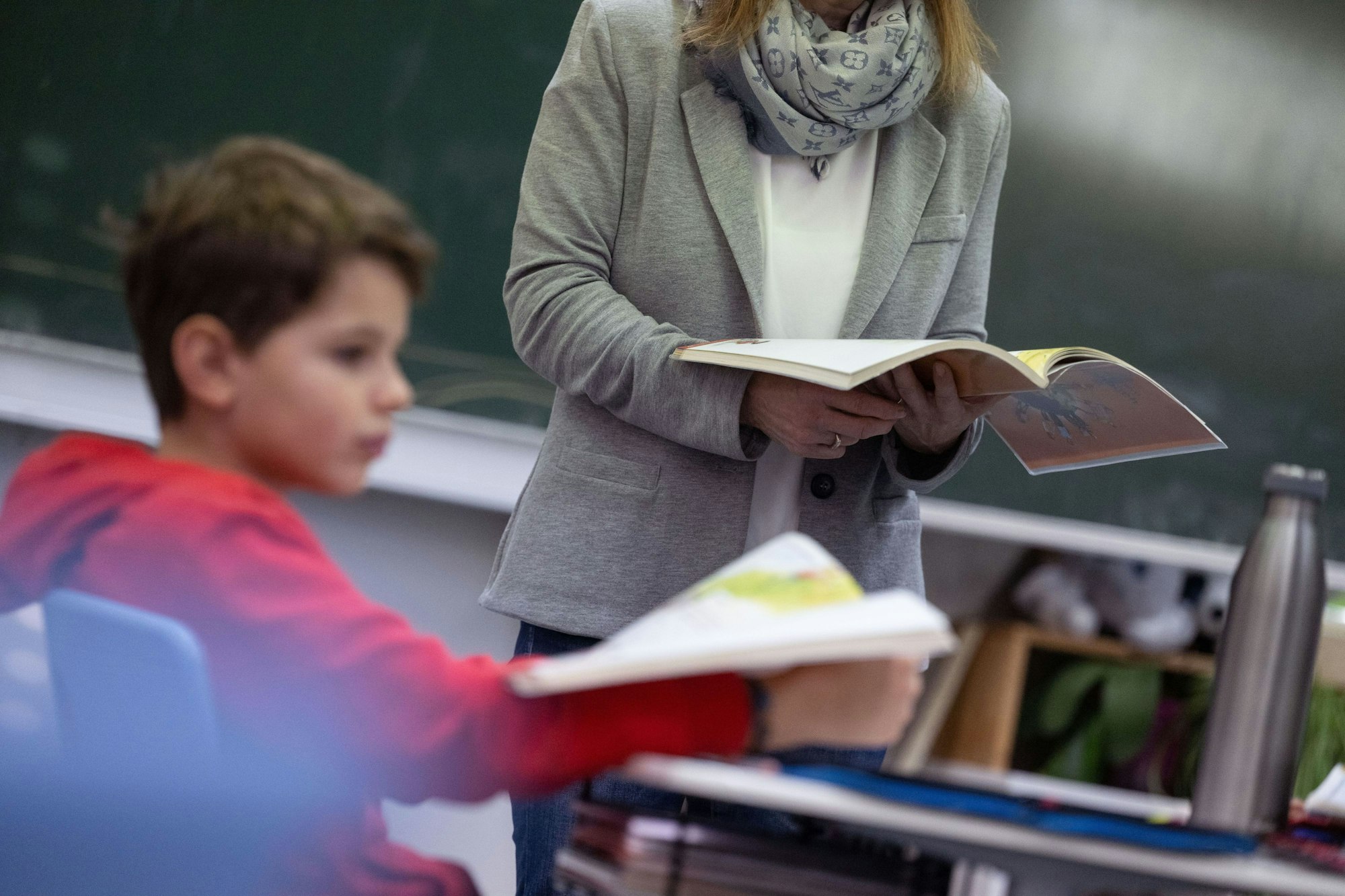 Ein Schüler einer vierten Klasse nimmt am Unterricht teil. Er trägt einen roten Pulli und hält ein Buch in der Hand. Vor ihm steht eine Lehrerin.