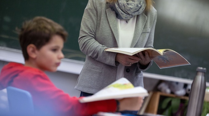 Ein Schüler einer vierten Klasse nimmt am Unterricht teil. Er trägt einen roten Pulli und hält ein Buch in der Hand. Vor ihm steht eine Lehrerin.&nbsp;
