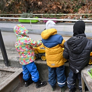 Man sieht drei Kinder, die auf einem Balkon spielen. Sie tragen Wintersachen und stehen mit dem Rücken zur Kamera.