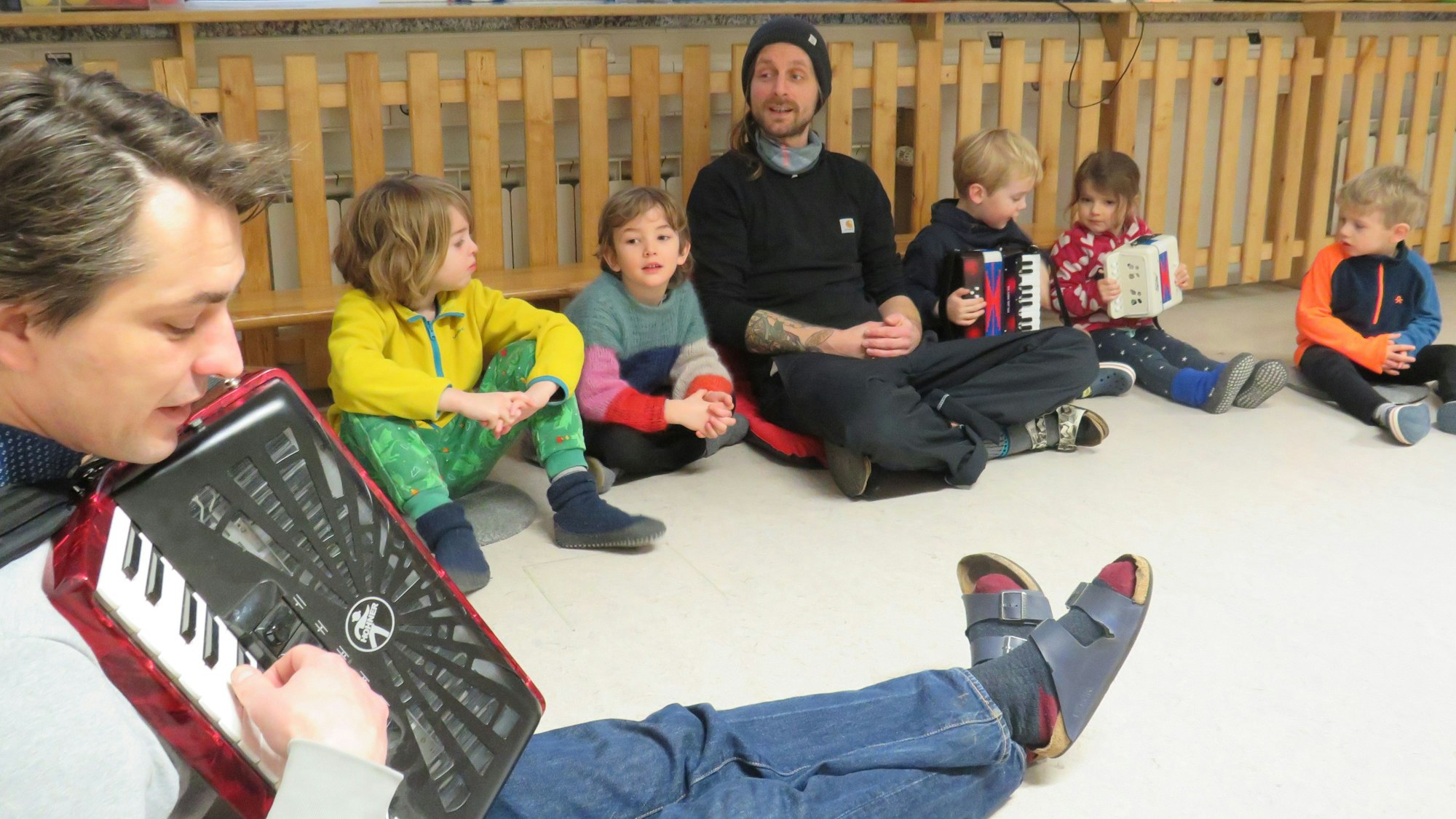 Kinder und zwei Erzieher sitzen auf dem Boden und singen gemeinsam. Ein Erzieher spielt auf einem Akkordeon.