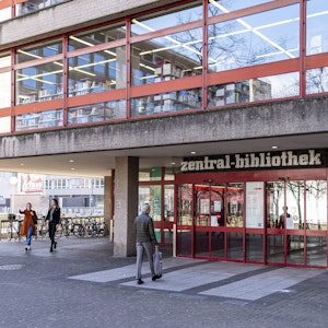 Das Foto zeigt den Eingang und einen Teil des verglasten ersten Stockwerks der Kölner Zentralbibliothek am Neumarkt. Passanten bewegen sich vor dem Gebäude.