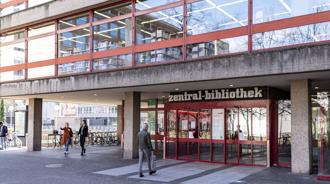 Das Foto zeigt den Eingang und einen Teil des verglasten ersten Stockwerks der Kölner Zentralbibliothek am Neumarkt. Passanten bewegen sich vor dem Gebäude.