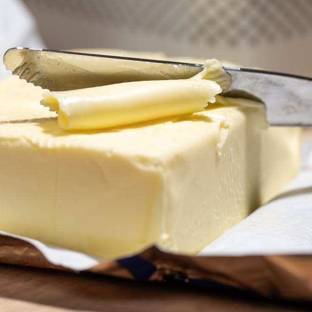 Ein Stück Butter liegt in der Verpackung auf dem Tisch, jemand nimmt mit dem Messer davon.