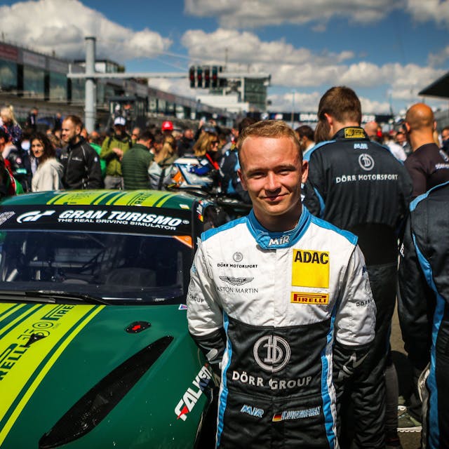 Moritz Wiskirchen steht beim 24-Stunden-Rennen 2022 vor einem Aston Martin. In der Boxengasse im Hintergrund sind viele Menschen.