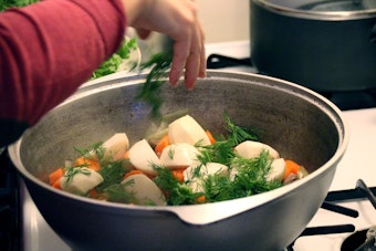 Eine Person wirft Zutaten für einen gekochten Salat mit Auberginen, Zwiebeln, Möhren, Tomaten, Dill und Koriander in eine Schüssel.