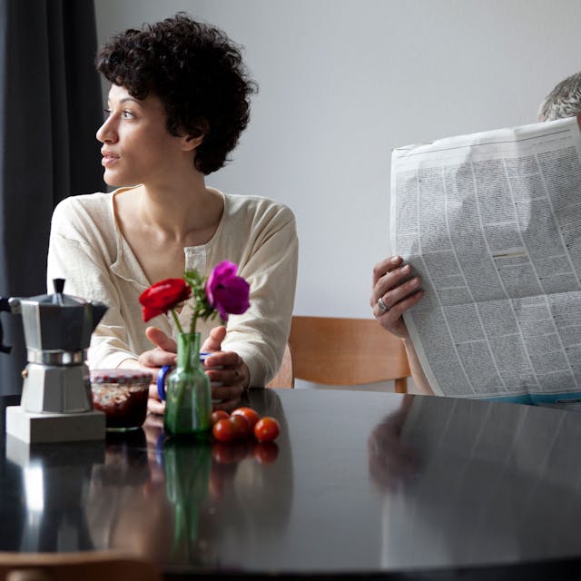 Eine Frau und ein Mann sitzen an einem Tisch. Die Frau schaut aus dem Fenster, der Mann in eine Zeitung.