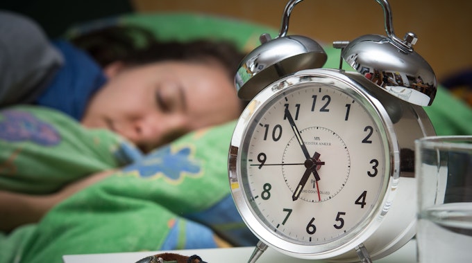 Eine Frau schläft im Bett, während ein Wecker, Wasserglas, Schmuck und ein Buch vor ihr auf dem Nachttischränkchen zu sehen sind.
