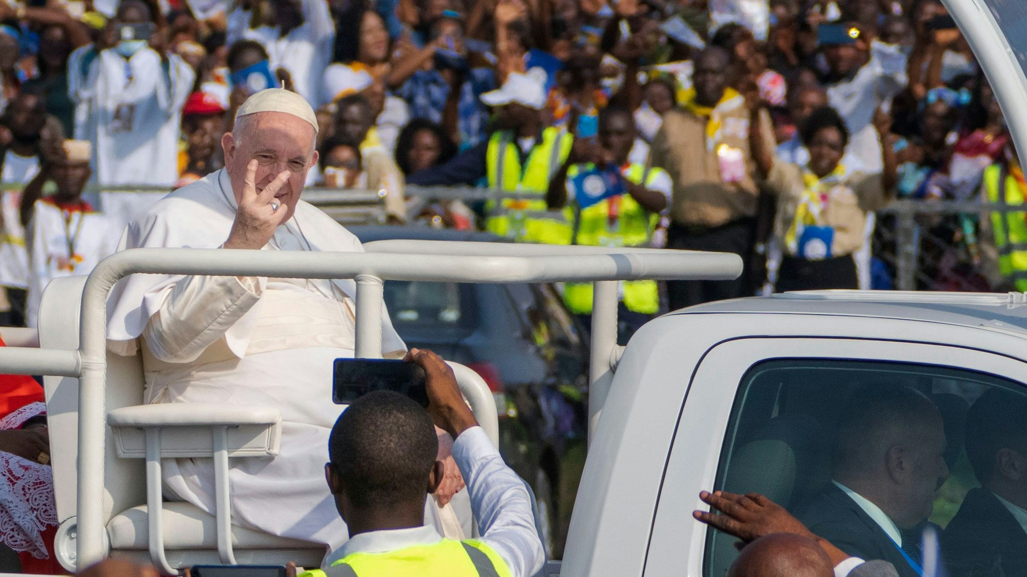 Papst Franziskus (M,l) sitzt bei seiner Ankunft auf dem Flughafen Ndolo in Kinshasa (Demokratische Republik Kongo) in einem offenen Fahrzeug und bildet mit den Fingern das V-Zeichen. Eine große Menge winkt ihm zu. Im Vordergrund fotografiert ein Mann in gelber Weste den Pontifex.