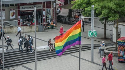 Im Juni 2021 weht auf dem Rathausvorplatz in Wiesdorf eine Regenbogenfahne. Passanten flanieren, Schüler stehen beieinander.