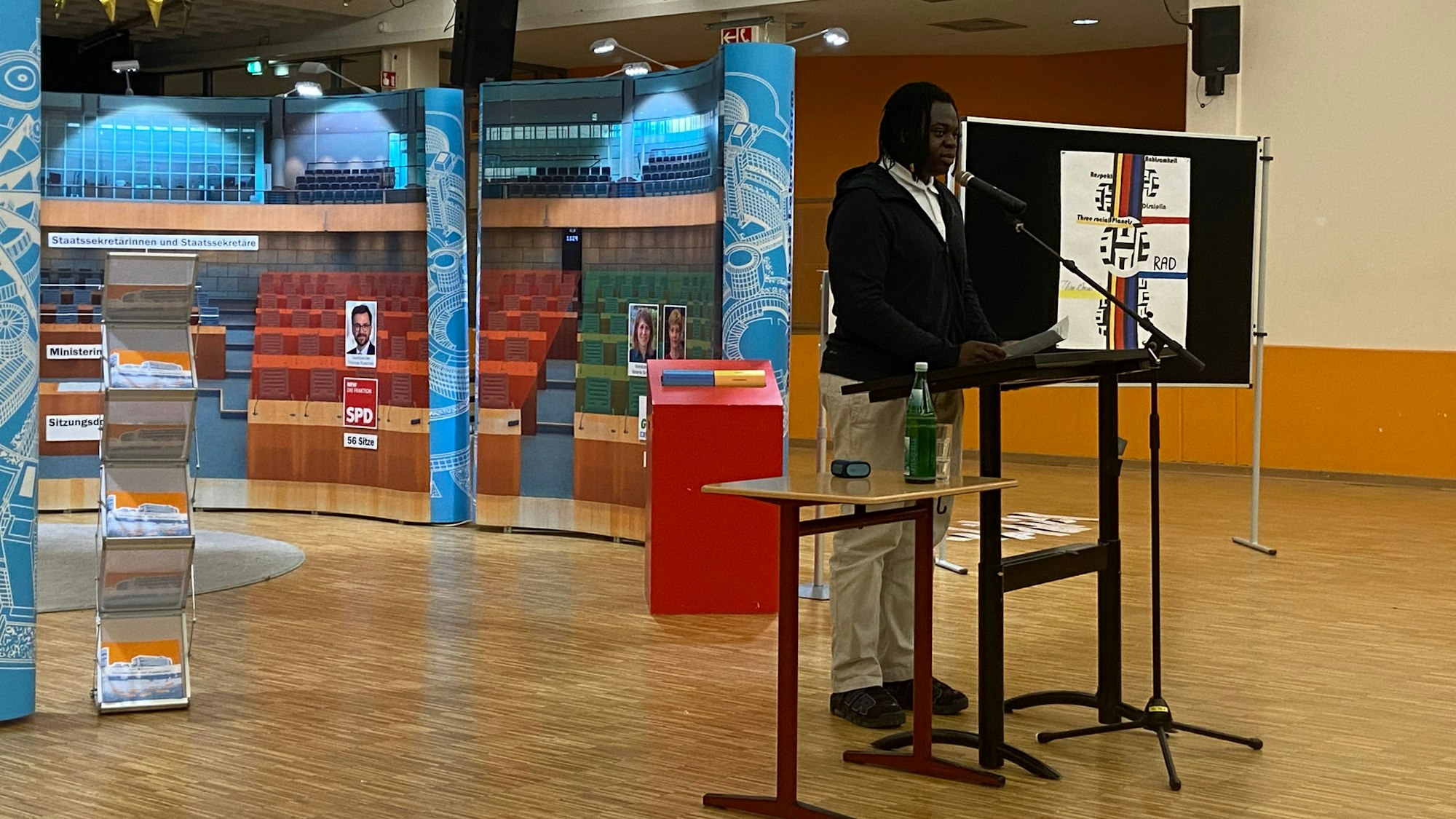 Ein Schüler steht an einem Mikrofon, hinter ihm ist eine Nachstellung des NRW-Landtags zu sehen.