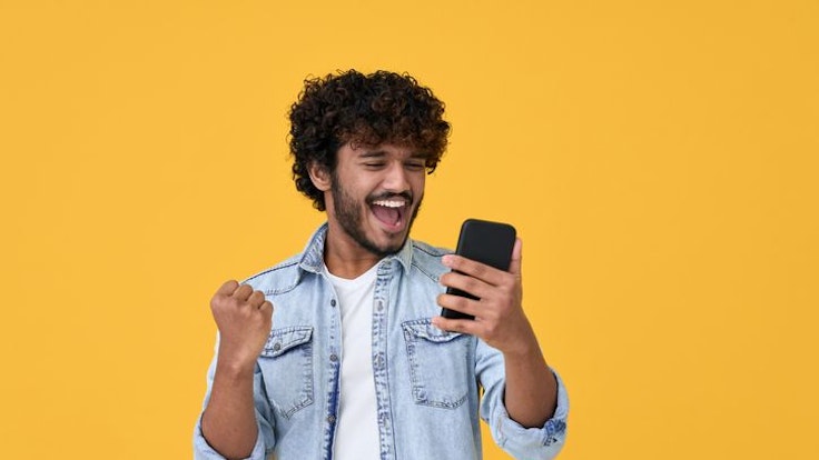 Aufgeregter junger Mann, der auf sein Smartphone blickt und sich freut, vor gelbem Hintergrund
