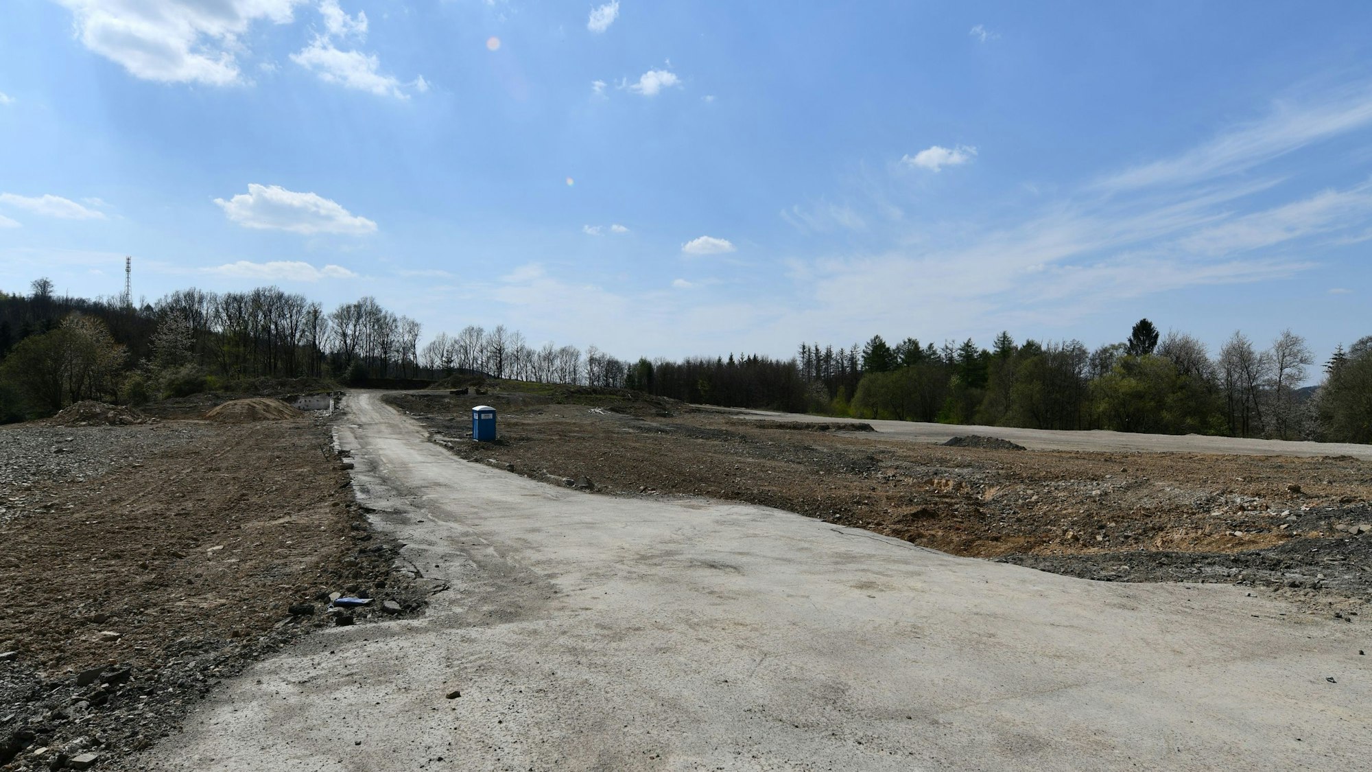 Ein leeres Feld aus Sand und Erde, in der Mitte ein Weg und ein Mülleimer. Im Hintergrund einige laublose Bäume vor strahlend blauem Himmel.