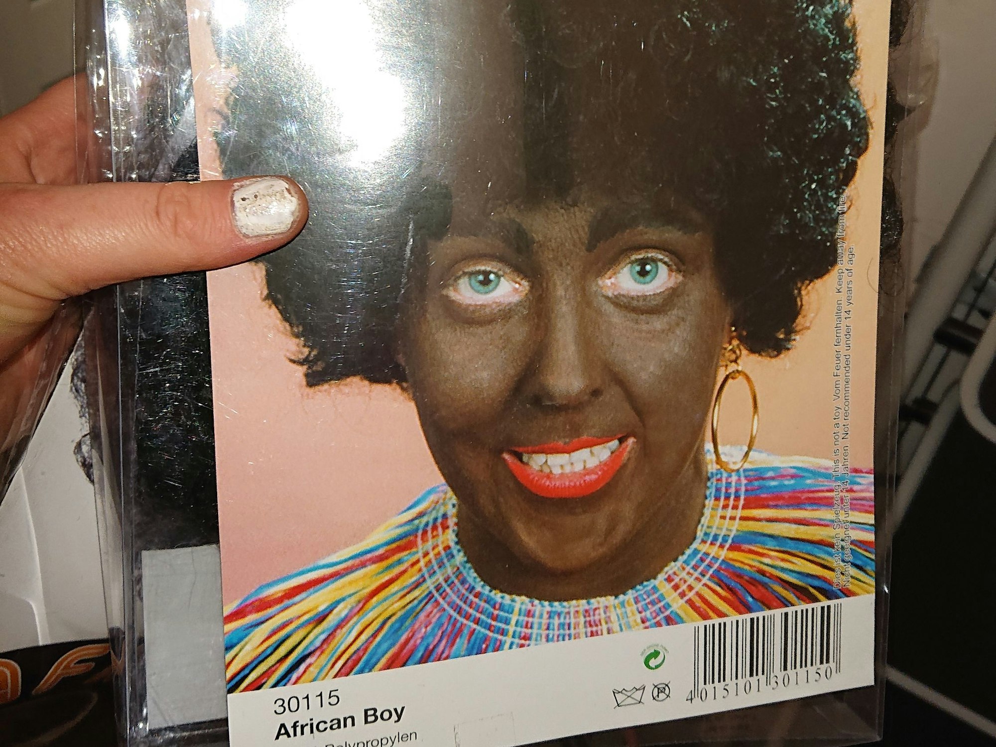 Eine schwarze Perücke als Karnevalskostüm mit der Verpackungsaufschrift „African Boy“. Auf dem Bild ist eine hellhäutige Frau abgebildet, die ihr Gesicht dunkelbraun geschminkt hat.