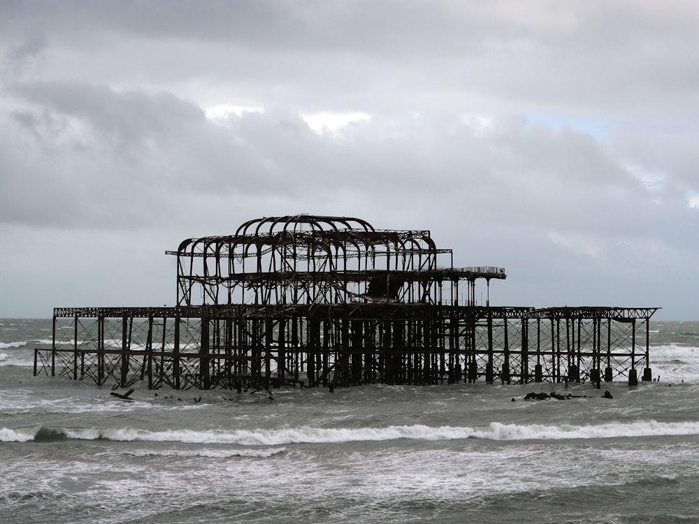 Das Foto von 2017 zeigt das Metallgestell des Brightoner Piers inmitten der Wellen, der Himmel ist grau.