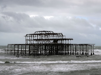 Das Foto von 2017 zeigt das Metallgestell des Brightoner Piers inmitten der Wellen, der Himmel ist grau.