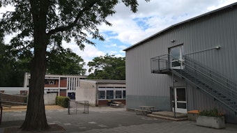 Auf einem Schulhof sind ein großer Baum und ein Container-Gebäude zu sehen, im Hintergrund zwei Flachbauten älteren Datums.