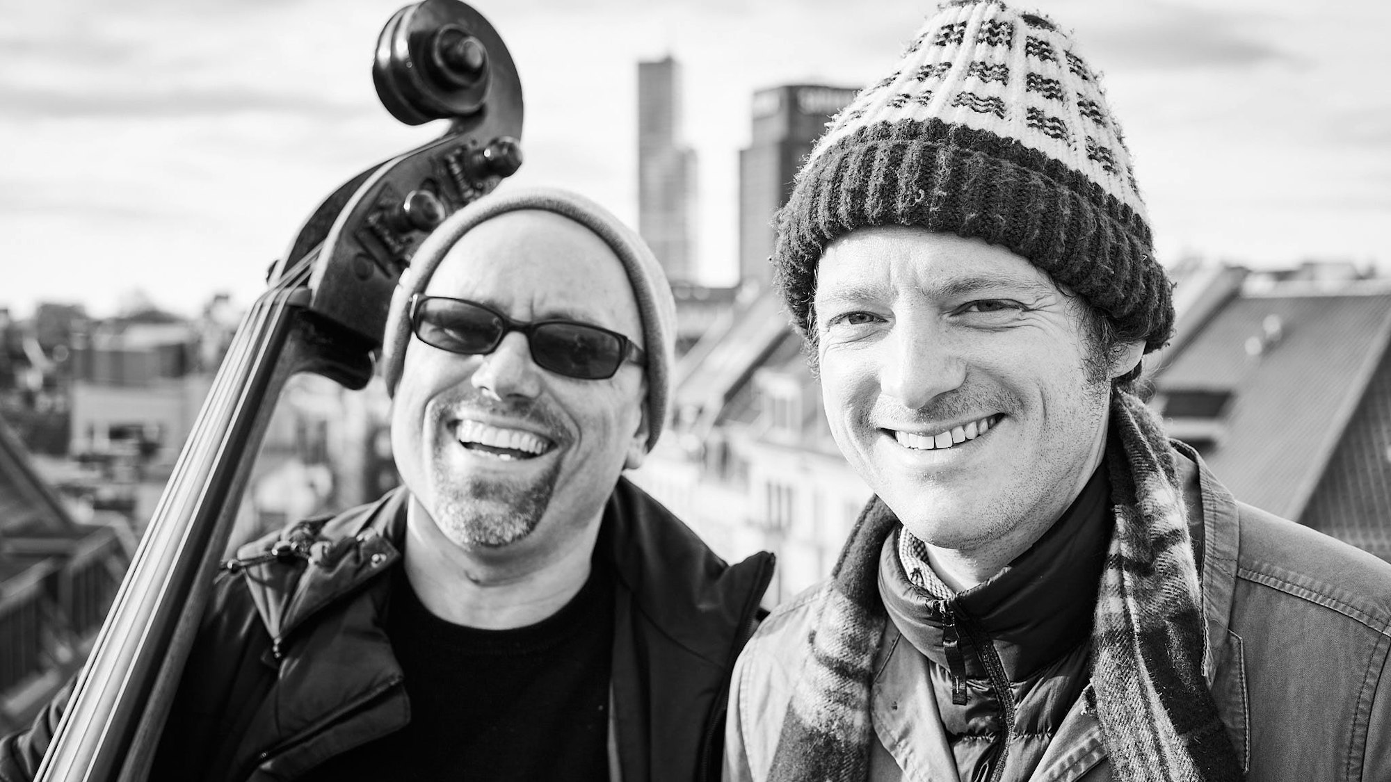 Die beiden Musiker Perfektomat alias Joscha Oetz und Retrogott alias Kurt Tallert vor der Kölner Skyline. Im Hintergrund erkennt man das Saturn-Hochhaus und den Mediapark. Oetz hält seinen Kontrabass.