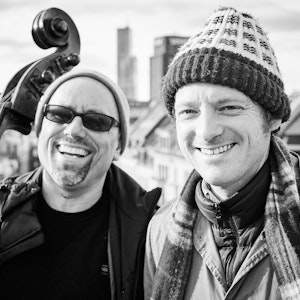 Die beiden Musiker Perfektomat alias Joscha Oetz und Retrogott alias Kurt Tallert vor der Kölner Skyline. Im Hintergrund erkennt man das Saturn-Hochhaus und den Mediapark. Oetz hält seinen Kontrabass.