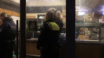 Hinter der Schaufensterscheibe eines Imbisses am Eigelstein sieht man uniformierte Beamtinnen und Beamte von Polizei und Zoll.
