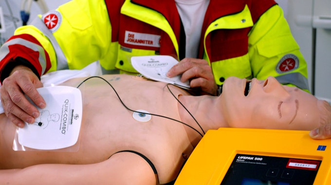 Ein Rettungssanitäter präsentiert einen Defibrillator an einer Puppe, die Elektroden-Pads liegen auf dem Torso.