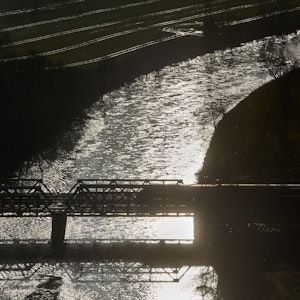 Ein Zug fährt im Gegenlicht über eine Brücke über das Flüsschen Lenne, in dessen Wellen sich die Morgensonne spiegelt.