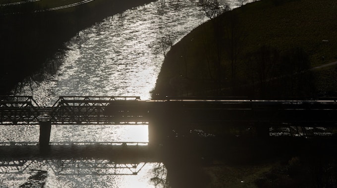 Ein Zug fährt im Gegenlicht über eine Brücke über das Flüsschen Lenne, in dessen Wellen sich die Morgensonne spiegelt.