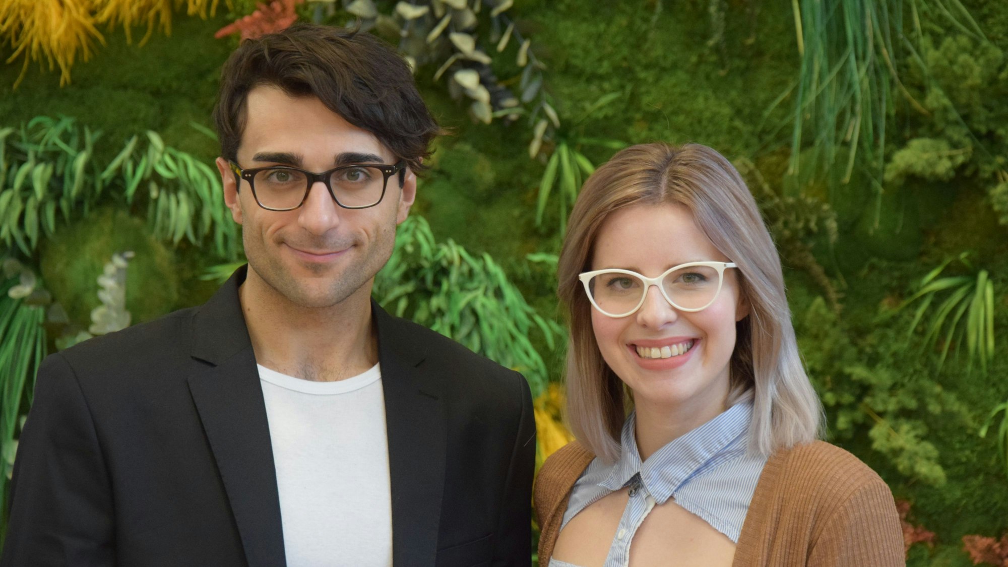 Deniz Tuzsus und Dana Pietralla lächeln in die Kamera. Sie stehen nebeneinander vor einer mit Pflanzen bedeckter Wand, die verschwommen ist.
