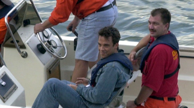 Robbie Williams sitzt in einem Boot der DLRG auf dem Rhein.