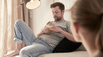 Eine Frau schaut einen Mann an, der mit etwas Abstand auf dem Sofa sitzt und etwas gelangweilt auf sein Handy blickt.