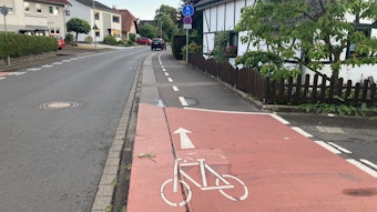 Über eine Kreuzung ist eine rote Markierung für einen Fahrradweg, dahiner folgt ein geteilter Fuß- und Radweg, auf dem auch Autos parken.