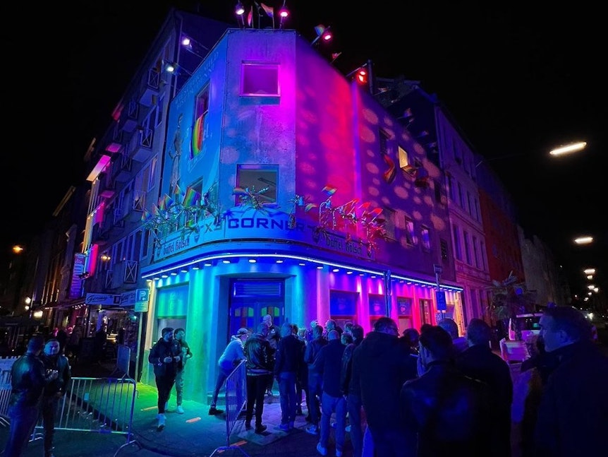 Zahlreiche Gäste stehen abends vor der in Regenbogenfarben beleuchteten ExCorner Bar.