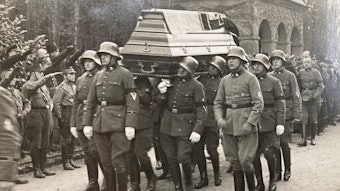 Uniformierte tragen den Sarg des Attentäters Hannes Miebach über den Melaten-Friedhof. Nationalsozialisten stehen Spalier und salutieren mit dem „Hitler-Gruß“.