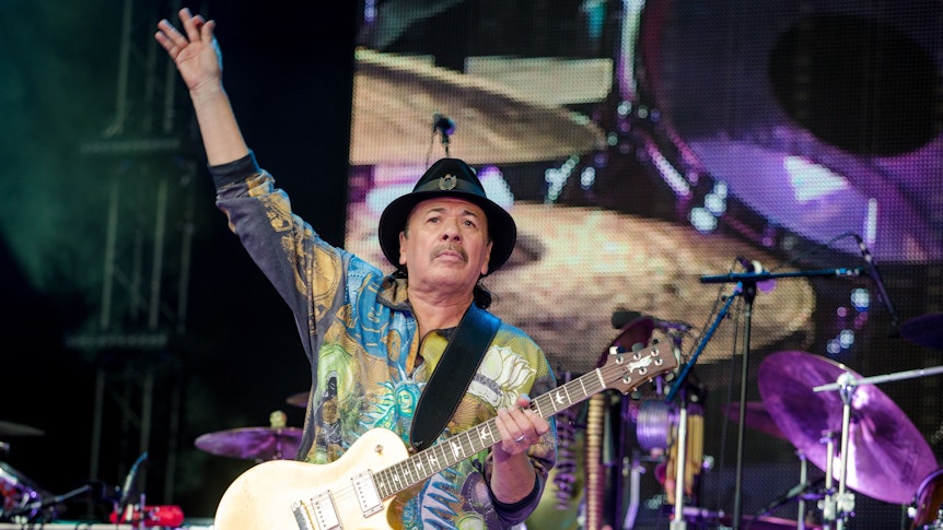 Das Foto aus dem Jahr 2016 zeigt den Gitarristen Carlos Santana. Er steht auf einer Bühne, hat einen Hut auf, ein buntes Shirt an und eine Gitarre umgehängt. Den rechten Arm streckt er nach oben, während er in die Menge schaut.