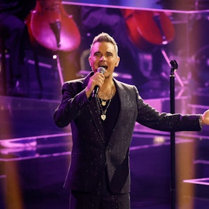 Robbie Williams steht in einem schwarzen Anzug auf der Bühne.
