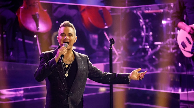 Robbie Williams steht in einem schwarzen Anzug auf der Bühne.