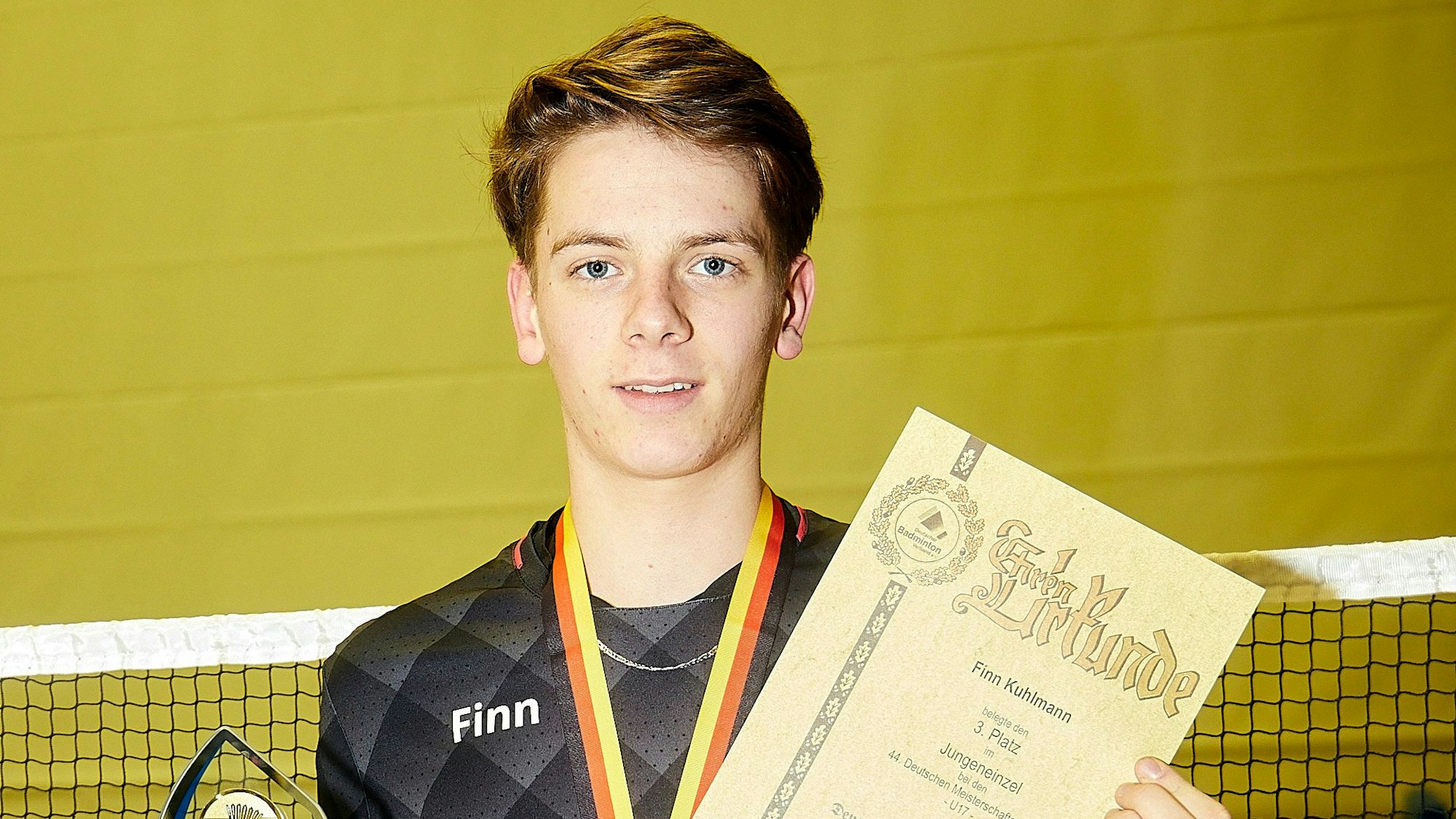 Finn posiert mit einer Urkunde und einem Pokal (der Deutschen Meisterschaften im Badminton). Er lächelt in die Kamera.