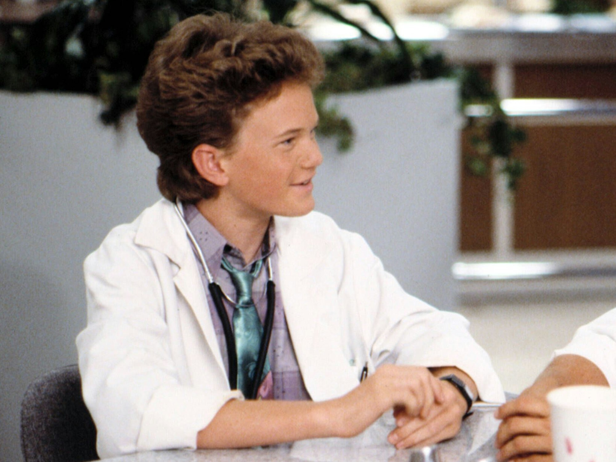 Der Kinderdarsteller Neil Patrick Harris sitzt in seiner Rolle als Dr. Douglas „Doogie“ Howser mit Arztkittel einer anderen Person gegenüber.