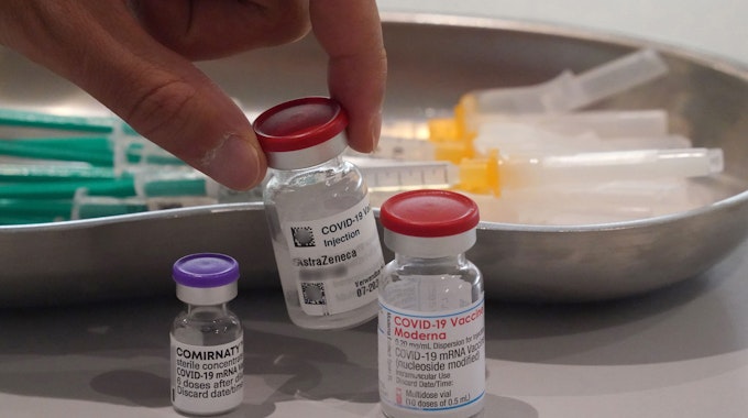 Ein Mitarbeiter eines Impfzentrums nimmt eine Impfdose vom Impfstoff Astrazeneca vom Tisch. Links im Bild steht eine Impfdose vom Impfstoff Biontech, rechts im Bild steht eine Impfdose vom Impfstoff Moderna.