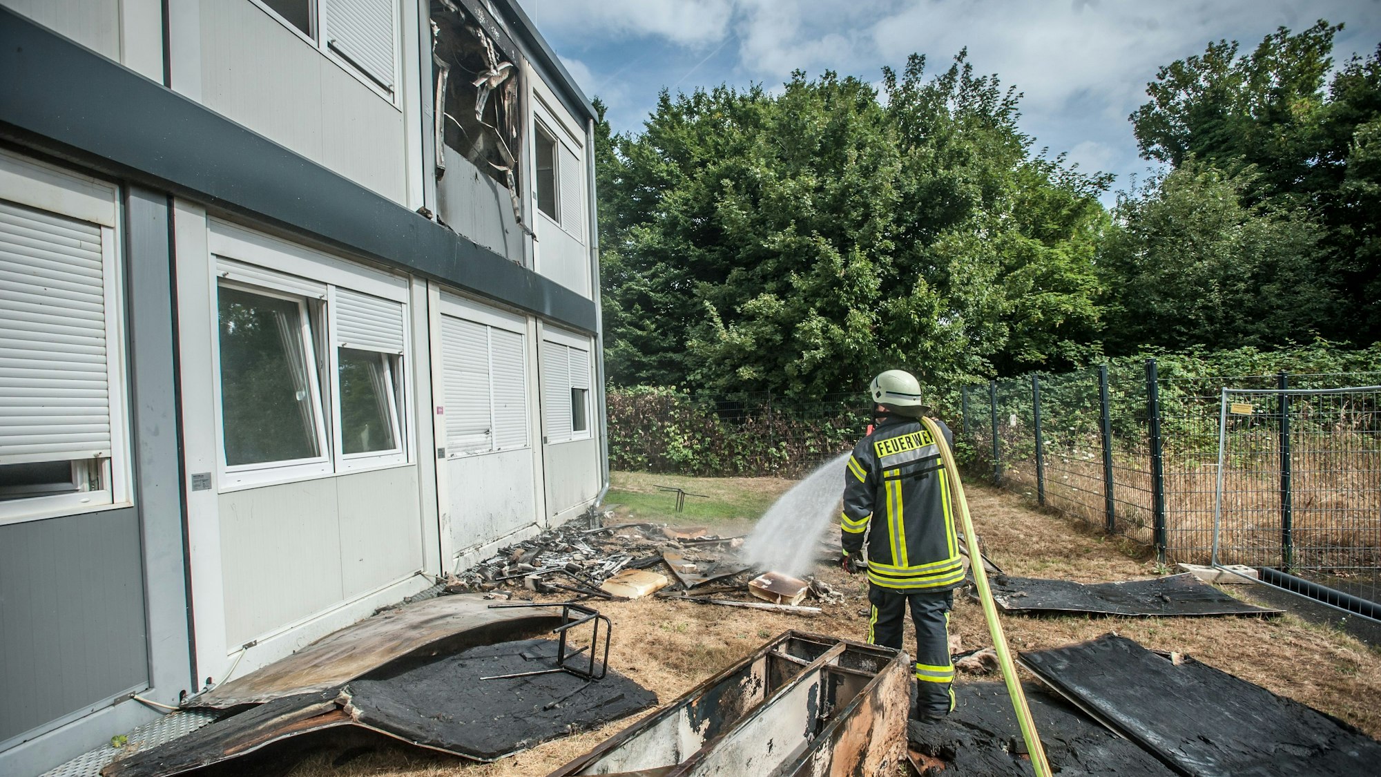 Ein Feuerwehrmann sprüht mit einem Schlauch Wasser auf verkohltes Material vor Wohncontainern.