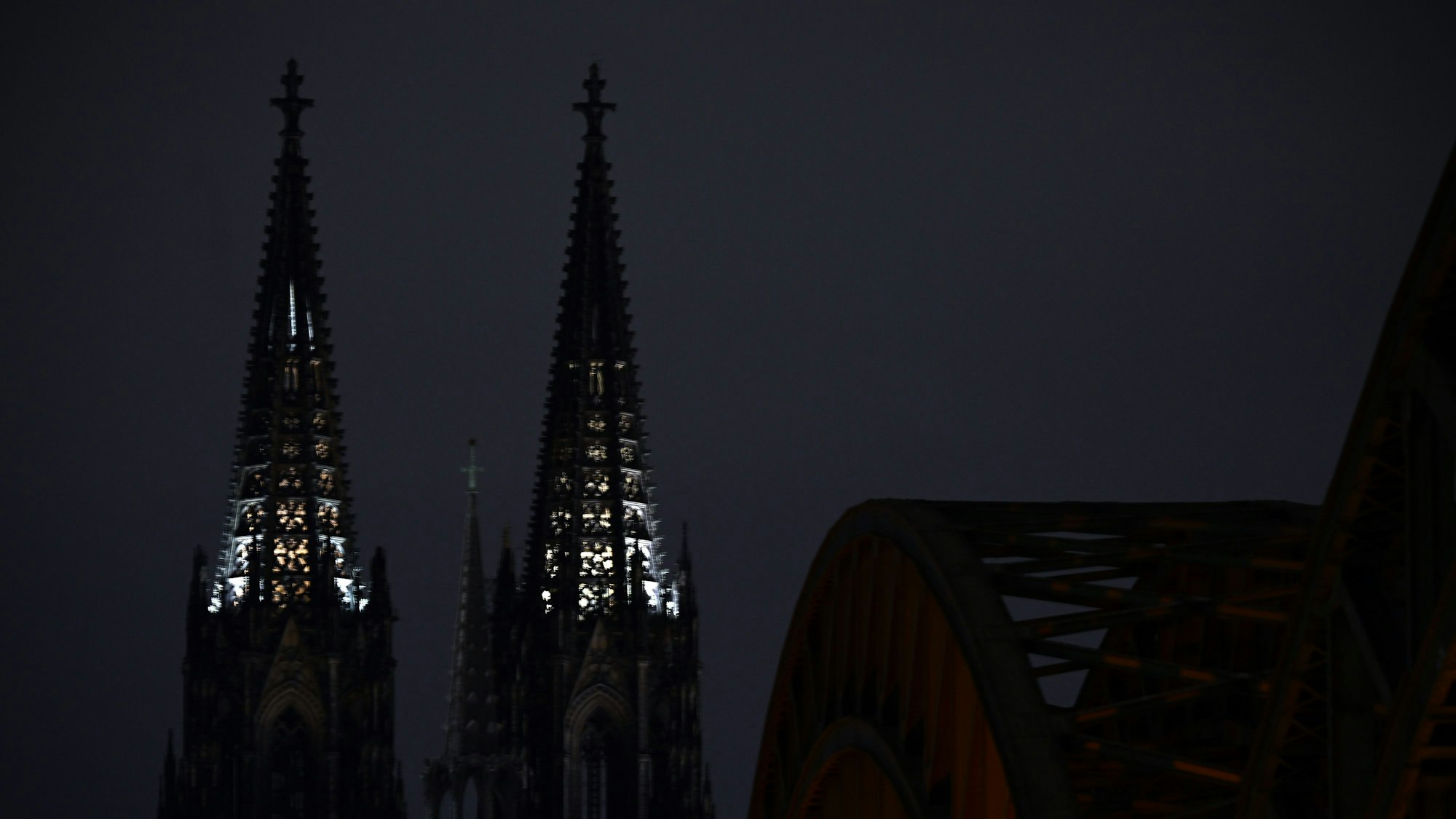 Vor nächtlichem Himmel heben sich die beiden Türme des Kölner Doms ab. Die Turmspitzen sind von innen beleuchtet. Rechts ist im Vordergrund die Hohenzollernbrücke zu sehen.