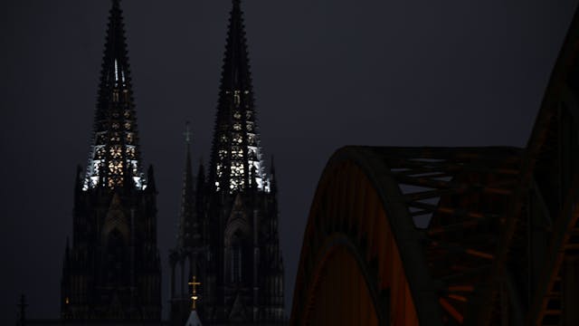 Vor nächtlichem Himmel heben sich die beiden Türme des Kölner Doms ab. Die Turmspitzen sind von innen beleuchtet. Rechts ist im Vordergrund die Hohenzollernbrücke zu sehen.