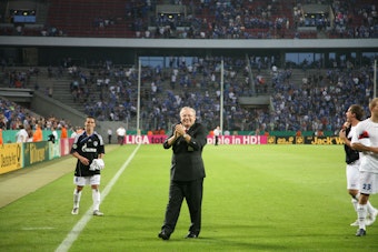 Franz-Josef Wernze hält seine Hände freudig in einem Fußball Stadion zusammen.