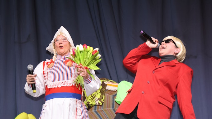 Zwei Frauen stehen auf der Bühne. Eine verkleidet als Frau Antje, mit bunten Tulpen in der Hand, die andere als Heino, der in ein Mikrofon singt.