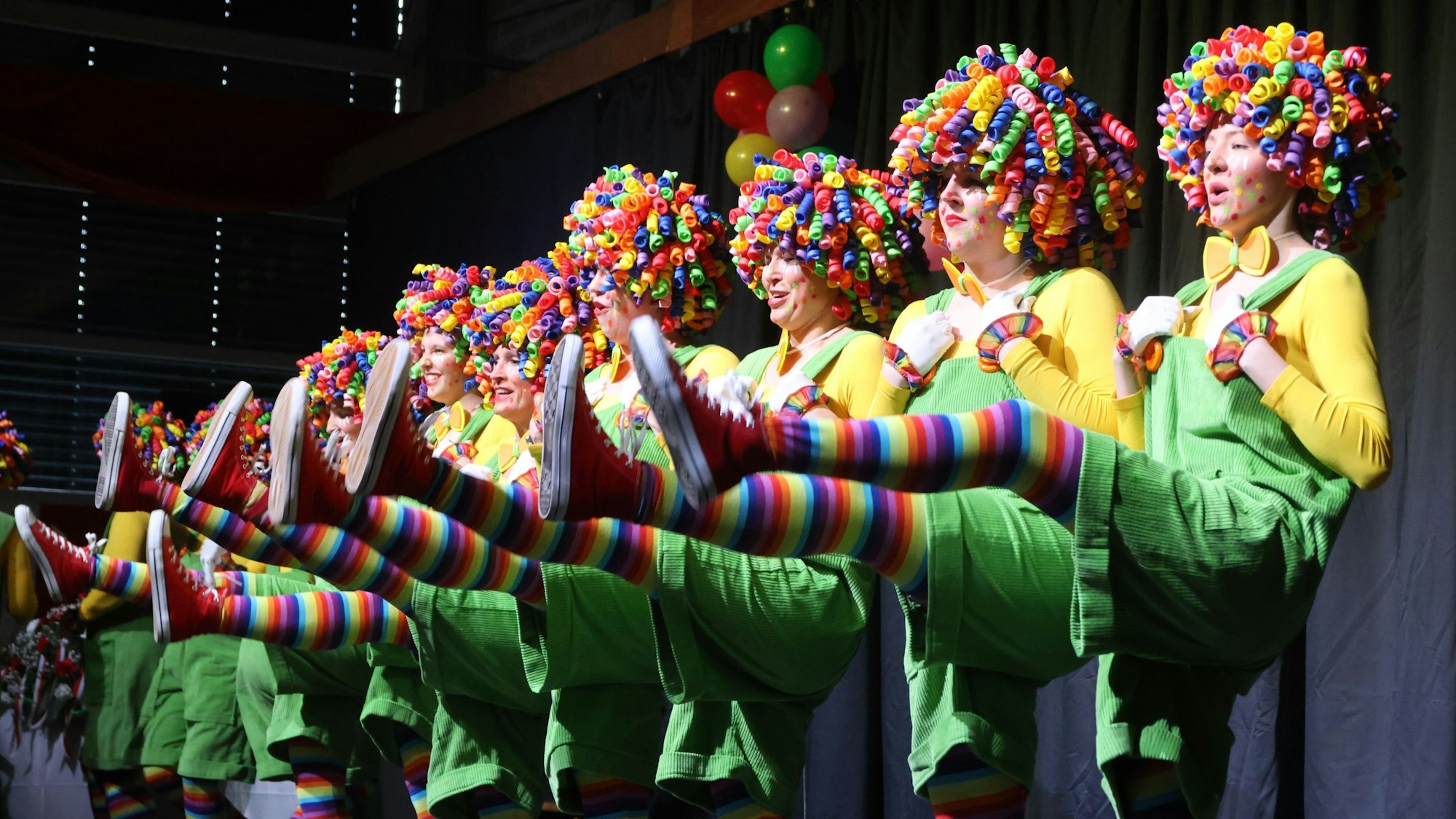 Mehrere Frauen, alle als Clowns mit bunten Perücken verkleidet, stehen tanzend in einer Reihe und schwingen jeweils ein Bein in die Höhe.